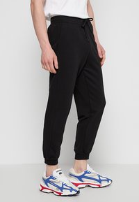 Zwarte joggingbroek met een relaxte pasvorm, elastische tailleband en boorden. Gecombineerd met witte sneakers met blauwe, rode en grijze accenten.