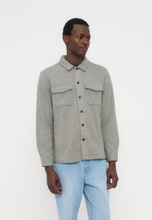 Άνδρας με ανοιχτογκρι πουκάμισο με κουμπιά και μπροστινές τσέπες με καπάκι, και ανοιχτό μπλε τζιν, στέκεται μπροστά σε απλό λευκό φόντο.