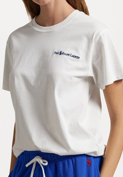 Balta medvilninė marškinė su apvaliu kaklu ir trumpomis rankovėmis, suteikiančios galimybes, su tamsiai mėlynu siuvinėjimu ant krūtinės. Lygus paviršius.