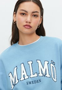Világoskék pulóver laza hímzéssel, amelyen a "MALMÖ" és a "SWEDEN" felirat tengerészkék és fehér színben szerepel, környakkal és puha tapintású anyaggal.