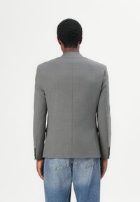 Grå skreddersydd blazer med strukturerte skuldre, enkelt vent, og knappeermer, sammen med lyseblå jeans, sett fra baksiden.