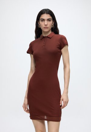 Woman with long dark hair wearing a short-sleeve, rust-colored fitted dress with a collar and button details, standing against a plain background.