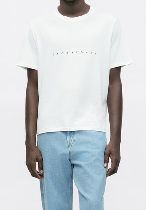 Person iført en hvid T-shirt med korte ærmer med teksten "JACK & JONES" og lyseblå jeans, stående mod en ensfarvet baggrund.