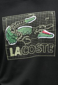 Μαύρη ζακέτα με γραφικά στοιχεία κεντημένα σε πράσινο και άσπρο, σχέδιο πλέγματος και το κείμενο "LACOSTE" με έντονες κίτρινες γραμμές.