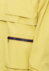 Casaco amarelo de exterior com um acabamento texturizado, apresentando um bolso com fecho éclair preto e detalhes coloridos em listras.