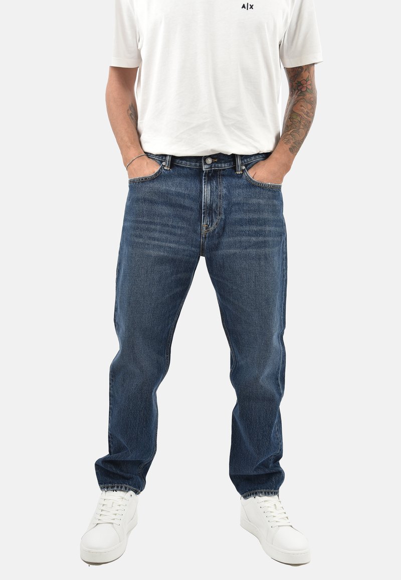 Jeans in denim blu scuro con un taglio classico, dotati di tasche frontali e una leggera scoloritura. Abbinati a sneakers bianche per un contrasto.