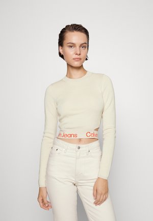 Maglione corto color crema con maniche lunghe e texture a coste, caratterizzato da un logo arancione Calvin Jeans sulla vita. Indossato con jeans chiari.