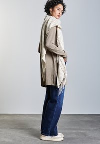 Beige långärmad topp, krämfärgad stickad sjal med fransar, mörkblå vida jeans och ljusa sneakers med beige detaljer. Enfärgad bakgrund.