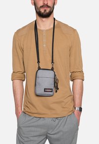 Borsa a tracolla grigia con chiusura a zip, cinturino regolabile e tasca frontale, realizzata in tessuto con una texture liscia. Presenta un logo del marchio.