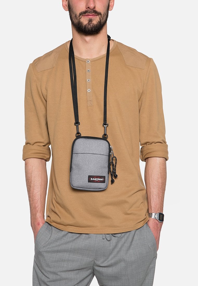 Borsa a tracolla grigia con chiusura a zip, cinturino regolabile e tasca frontale, realizzata in tessuto con una texture liscia. Presenta un logo del marchio.
