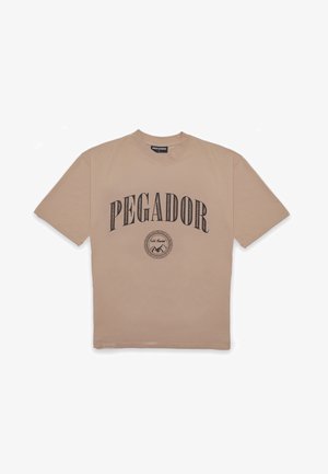 Camiseta de algodón beige con mangas cortas, con la leyenda "PEGADOR" impresa en letras negras en negrita y un diseño gráfico circular debajo.