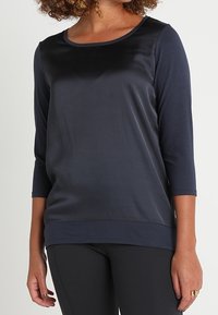 Frau, die ein navy-blaues, langärmliges Satin-Front-Shirt mit rundem Ausschnitt und schwarze enganliegende Hosen trägt.