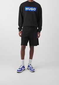 Sweatshirt noir avec un logo "HUGO" bleu, associé à des shorts noirs. Porté avec des chaussettes blanches et des baskets bleu et blanc. Design simple.