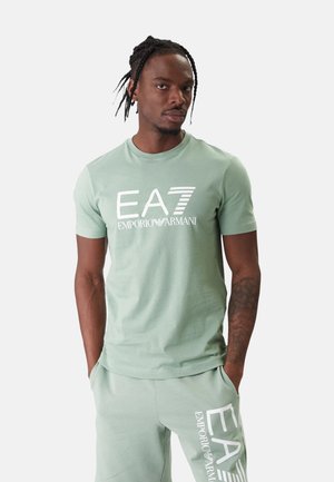 Uomo con capelli intrecciati che indossa una t-shirt verde chiaro EA7 Emporio Armani e pantaloncini abbinati con le mani in tasca su sfondo uniforme.