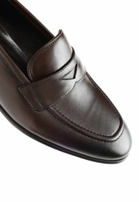 Mocassin en cuir marron avec un bout rond, des accents cousus et une bande décorative sur le devant. Texture lisse et design élégant.