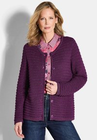 Lila gerippter Cardigan mit rundem Ausschnitt, strukturierter Oberfläche, fünf Knöpfen und langen Ärmeln, über einer gemusterten pinken Bluse getragen.