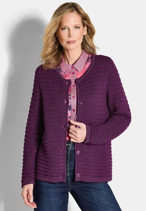 Lila gerippter Cardigan mit rundem Ausschnitt, strukturierter Oberfläche, fünf Knöpfen und langen Ärmeln, über einer gemusterten pinken Bluse getragen.