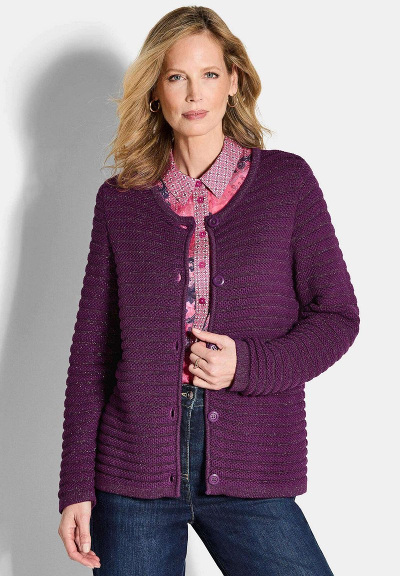 Lila gerippter Cardigan mit rundem Ausschnitt, strukturierter Oberfläche, fünf Knöpfen und langen Ärmeln, über einer gemusterten pinken Bluse getragen.