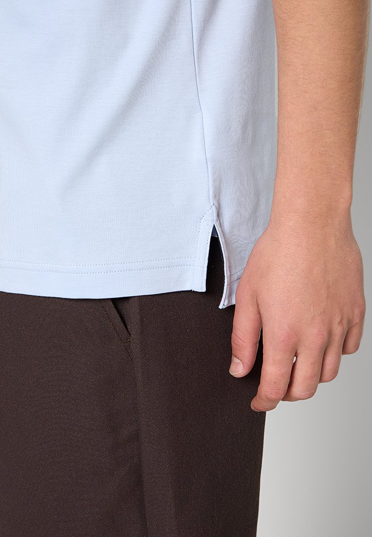 Detailní záběr na ruku osoby odpočívající podél těla, oblečenou v světle modré košili s bočním rozparkem a tmavě hnědých kalhotách s kapsou.