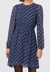 Modrá šaty s vzorem, dlouhými průhlednými rukávy, shromážděným pasem a splývavou sukní, s geometrickým designem v světlé modré na tmavém pozadí.