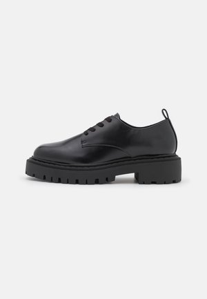Chaussure noire en cuir à lacets avec semelle épaisse et robuste et languette de traction sur fond blanc, vue de côté.