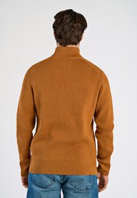 Maglione rigato marrone con colletto alto, maniche lunghe e taglio aderente. La texture è morbida e presenta un'etichetta logo laterale.