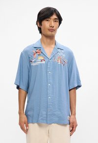 Γαλάζιο κοντομάνικο πουκάμισο με κεντημένα μοτίβα ήλιου, κυμάτων και λουλουδιών. Διαθέτει κουμπιά μπροστά και άνετη εφαρμογή.