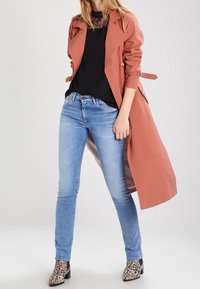 Trench-coat rosé avec ceinture à la taille, blouse noire, jean skinny bleu clair et bottines à motif animalier. Tissu lisse et coupe ajustée.