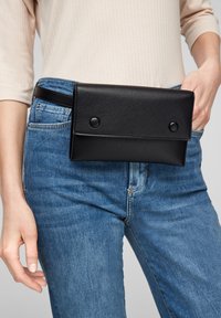 Pochette en cuir noir à finition texturée, avec deux boutons-pression, portée sur une ceinture noire fine au-dessus d'un jean bleu.