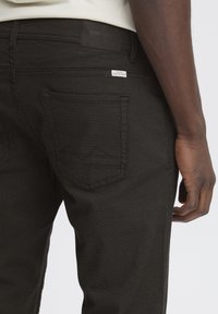 Črne jeans hlače z teksturiranim zaključkom, vidnimi zadnjimi žepi s šivanimi detajli in blagovno oznako na pasu.