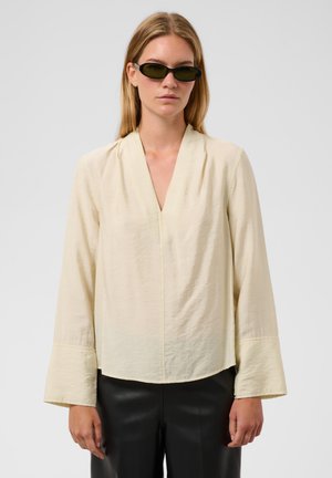 Vrouw met zwarte smalle zonnebril, crème lange-mouwen blouse met brede manchetten en zwarte broek, staand tegen een effen achtergrond.