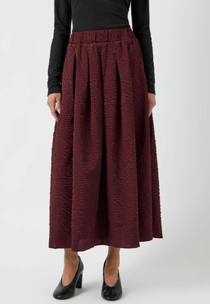 Žena nosi teksturiranu bordo midi suknju sa naborima i crni top s dugim rukavima, upareno s crnim cipelama na visoku petu.