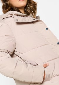 Doudoune beige clair avec fermeture éclair et pression, dotée d'une capuche, d'un tissu texturé et d'une poche latérale.