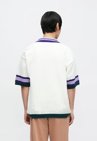 Biała dzianinowa koszulka polo z teksturowanym wzorem, krótkimi rękawami z fioletowo-zielonymi paskami oraz prążkowanym kołnierzykiem i mankietami.