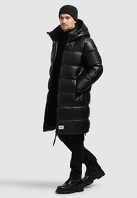 Cappotto imbottito nero con finitura lucida, dotato di cappuccio, chiusura con zip e un accento del logo. Indossato sopra un dolcevita nero e pantaloni.