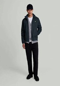 Mörkgrön jacka med krage, grå stickad cardigan under, svarta byxor och en svart keps. Enkelt design, inga synliga mönster.