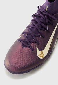 Chaussure de football violette avec une surface texturée, un Swoosh Nike blanc bien en vue, et un design de lacets complexe avec des accents dorés.