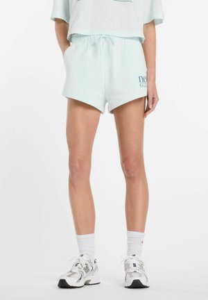 Kvinde iført lyseblå afslappede shorts med snøre i taljen, hvide crew-sokker og hvide New Balance sneakers, stående med hænderne i lommerne.