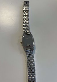 Orologio da polso in metallo argentato con quadrante rettangolare. Caratterizzato da una superficie lucida e liscia, con un bracciale a maglie in metallo e un piccolo corona laterale.