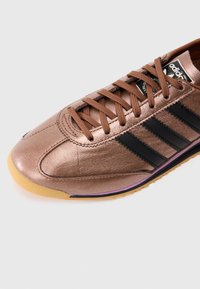 Χάλκινο μεταλλικό αθλητικό παπούτσι με μαύρες ρίγες, που διαθέτει καφέ κορδόνια, στρογγυλή μύτη και υφή στην επάνω επιφάνεια. Σόλα από καουτσούκ με μοβ λεπτομέρεια.