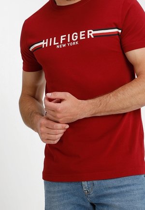 T-shirt rouge à manches courtes avec le texte "HILFIGER NEW YORK" et des détails rayés blancs, rouges et noirs sur la poitrine, porté par une personne.