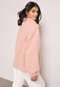 Šviesiai rožinė, pūkuota vilnos striukė su klasikiniu apykakle, nuleistais pečiais ir elastingomis rankovėmis; per didelis modelis.