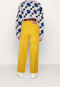 Zkrácený svetr s vícebarevným kostkovaným vzorem na levandulovém podkladu; spárovaný se žlutými manšestrovými kalhotami širokého střihu a bílými teniskami.