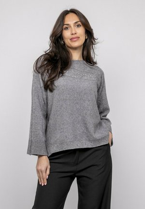 Femme aux longs cheveux noirs portant un pull gris en maille avec des détails subtils en perles et un pantalon noir, debout devant un fond clair uni.