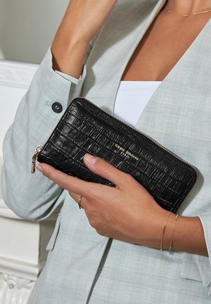 Zwarte portemonnee met krokodillenstructuur, met rits, vastgehouden in een hand, voorzien van gouden hardware en een subtiele merklabel, tegen een lichtgrijze blazer.