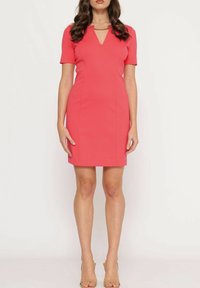 Žena na sobě má korálově růžové krátké šaty ke kolenům s výstřihem do V a zlatým řetízkem, které doplňuje béžovými otevřenými sandály na podpatku.