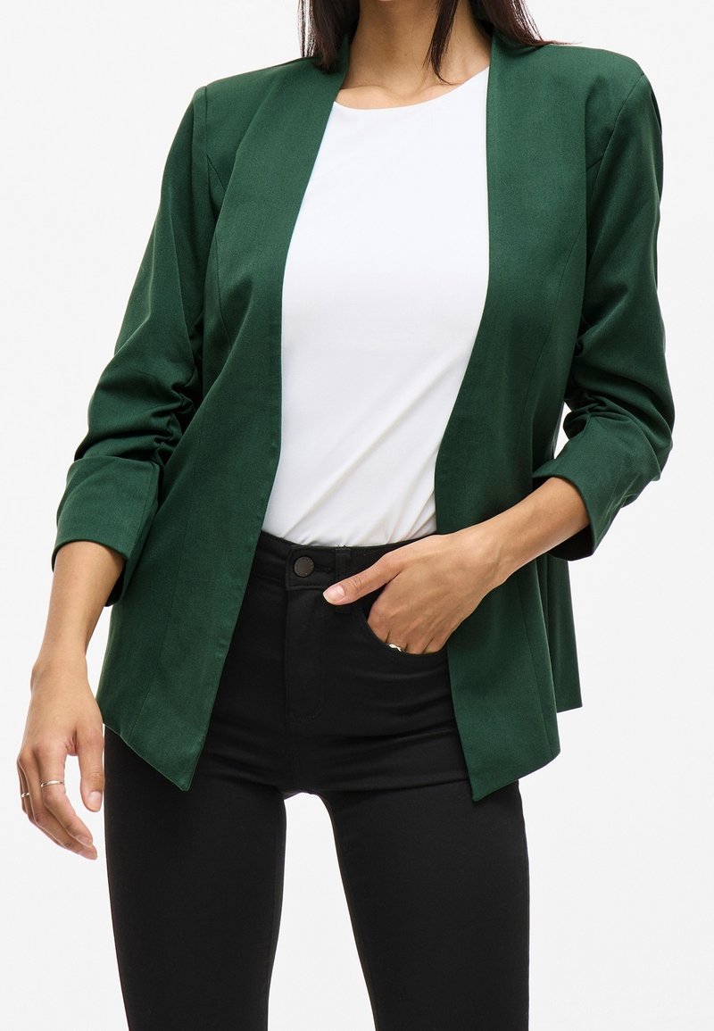 Femme portant un blazer vert foncé sur un haut blanc et un pantalon noir, main partiellement dans la poche, sur un fond blanc uni.