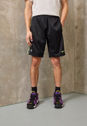 Osoba w czarnych szortach Nike z neonowymi akcentami oraz czarnych, fioletowo-żółtych sneakersach stoi na tle szarej betonowej ściany.