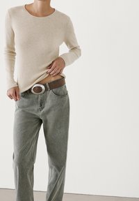 Beige geribbelde top met lange mouwen gecombineerd met grijze spijkerbroek. De outfit wordt geaccentueerd met een bruine riem met een zilveren ovale gesp, wat een casual stijl benadrukt.