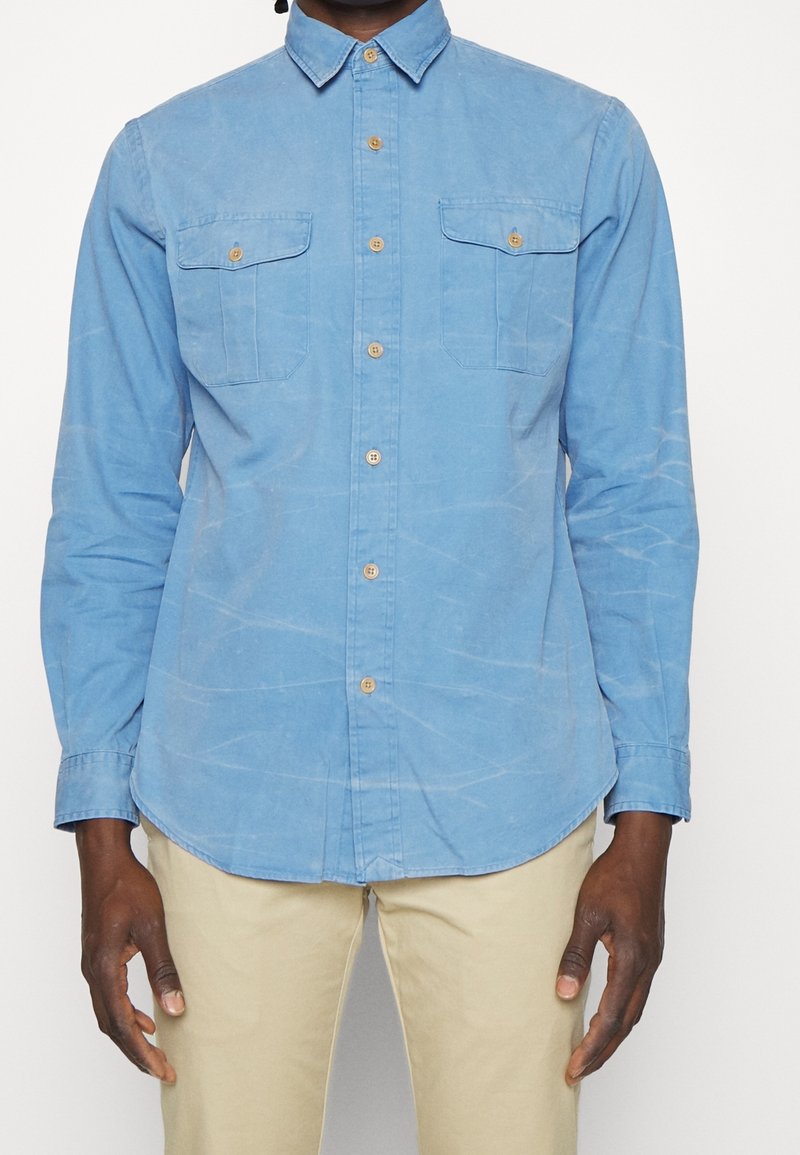 Light blue button-up shirt with a spread collar, two chest pockets, and long sleeves. Made of a textured fabric with a slightly wrinkled appearance.